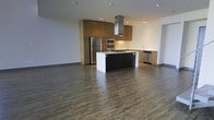This contemporary two-floor space is defined by clean lines, modern finishes, and a thoughtfully designed open layout. The main level showcases a striking open-concept kitchen that serves as the heart of the space. A large center island with a dark base and bright white countertop provides both a functional workspace and a natural gathering point. Warm wood-toned cabinetry lines the walls, offering generous storage, while a sleek stainless steel range hood anchors the cooking area with an industrial-modern aesthetic. High-end stainless steel appliances, including a full-size refrigerator, built-in wall ovens, a gas cooktop, and a dishwasher, complete the well-appointed kitchen. Recessed lighting throughout the ceiling ensures bright, even illumination across the open floor plan. Rich, dark hardwood-style flooring flows continuously through the space, grounding the interior with warmth and texture. The open plan living area extends generously from the kitchen, offering flexible use of the space. Clean, neutral-toned walls keep the atmosphere light and versatile. The two-floor configuration provides distinct zones for living and private use, making this a well-structured and functional space suited for a variety of needs.