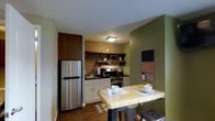 This single-floor space offers a thoughtfully designed kitchen and dining area that maximizes functionality within a compact footprint. The kitchen is anchored by a striking exposed brick backsplash, which adds texture and a warm, industrial character to the space. White lower cabinetry provides clean storage and contrasts effectively with the dark countertops, while a tall, wood-paneled upper cabinet offers additional storage alongside open shelving that displays small appliances and kitchenware. Stainless steel appliances, including a full-size refrigerator, a gas range, and a microwave, complete the kitchen's practical setup. Dark hardwood flooring runs throughout, grounding the space with warmth. An accent wall painted in deep olive green defines the dining area, where a live-edge wood table is mounted to the wall, paired with industrial-style metal stools. A wall-mounted television sits above, making this corner a versatile spot for both dining and relaxing. A flush-mount ceiling light provides functional illumination. Overall, the space balances rustic and modern elements cohesively.