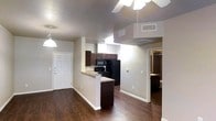 This single-floor residential space features an open-concept design that seamlessly connects the living area and kitchen, creating a functional and cohesive layout. The flooring throughout is a rich, dark hardwood that adds warmth and contrast to the neutral beige walls and white baseboards.

The kitchen is outfitted with dark espresso cabinetry, a granite-topped peninsula that doubles as a breakfast bar, and black appliances including a refrigerator, range, and microwave. A fluorescent light fixture illuminates the kitchen workspace, while a ceiling fan with an integrated light fixture serves the living area. A pendant light near the entry door adds additional ambient lighting.

The front entry door is solid white with a multi-lock setup, providing a secure and clean entryway directly into the open living space. A hallway visible toward the back of the unit leads to what appears to be a bedroom and bathroom, with a glimpse of a closet visible through the doorway.

HVAC ventilation is provided through ceiling-mounted vents, and the layout benefits from a straightforward, efficient flow between spaces. Overall, the unit presents a clean, modern aesthetic defined by its dark wood tones, neutral palette, and practical open floor plan.
