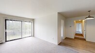 This single-floor unit offers a clean, functional layout with a neutral color palette throughout. The living area is a notable highlight, featuring soft carpeting underfoot and a large sliding glass door dressed with vertical blinds, which allows abundant natural light to fill the room while providing views of the surrounding greenery. A baseboard heating system is present, and the space connects smoothly to additional rooms via an adjoining hallway with wood-look flooring.

The kitchen is efficiently designed with gray cabinetry on both upper and lower sections, white countertops, and a double stainless steel sink positioned beneath a window with vertical blinds. A white electric coil-top range with an overhead exhaust hood completes the cooking area, and the wood-look flooring carries throughout this space.

The bedrooms are carpeted and well-proportioned, each featuring windows with vertical blinds that bring in natural light. One bedroom includes direct access to a full bathroom and a closet, offering added convenience. The bathroom is appointed with a full soaking tub and shower combination, a toilet, vanity, and tile flooring, with baseboard heating rounding out the practical amenities of this straightforward and functional unit.