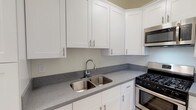 Located in the desirable Hollywood Hills neighborhood of Los Angeles, this single-floor residence at 1800 El Cerrito Pl offers a clean, updated interior with thoughtfully maintained details throughout. The kitchen is a standout feature, outfitted with crisp white shaker-style cabinetry, grey speckled stone countertops, a stainless steel double-basin sink, and stainless steel appliances including a gas range with oven and an over-the-range microwave. Stone tile flooring anchors the kitchen space with a warm, neutral tone. The main living area is bright and open, featuring grey-toned hardwood-style flooring, crown molding, a ceiling fan, and windows that allow natural light to filter in alongside views of surrounding greenery. An arched doorway adds a classic architectural charm to the space. The bedroom continues the hardwood-style flooring and crown molding, with a built-in closet and ample natural light from a window with blinds. A narrow hallway features a built-in white vanity shelf with a mirror, leading to additional storage and a walk-in closet area. The bathroom is finished with stone tile flooring, a pedestal sink, and a full tub-shower combination, maintaining a bright, clean aesthetic throughout.