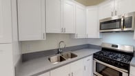 Located in the desirable Hollywood Hills neighborhood of Los Angeles, this single-floor residence at 1800 El Cerrito Pl offers a clean, updated interior with thoughtfully maintained details throughout. The kitchen is a standout feature, outfitted with crisp white shaker-style cabinetry, grey speckled stone countertops, a stainless steel double-basin sink, and stainless steel appliances including a gas range with oven and an over-the-range microwave. Stone tile flooring anchors the kitchen space with a warm, neutral tone. The main living area is bright and open, featuring grey-toned hardwood-style flooring, crown molding, a ceiling fan, and windows that allow natural light to filter in alongside views of surrounding greenery. An arched doorway adds a classic architectural charm to the space. The bedroom continues the hardwood-style flooring and crown molding, with a built-in closet and ample natural light from a window with blinds. A narrow hallway features a built-in white vanity shelf with a mirror, leading to additional storage and a walk-in closet area. The bathroom is finished with stone tile flooring, a pedestal sink, and a full tub-shower combination, maintaining a bright, clean aesthetic throughout.
