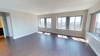 This single-floor space is characterized by its clean, open layout and modern finishes. Dark hardwood flooring extends throughout the main area, providing a rich contrast against the light neutral-toned walls and white textured ceiling. Three generously sized windows line the exterior wall, flooding the interior with abundant natural light and framing impressive elevated views of the surrounding cityscape, including a dynamic urban skyline with green spaces visible in the distance. The windows are equipped with horizontal blinds, offering adjustable light control while maintaining the connection to the outdoors. The textured white ceiling adds subtle visual interest overhead, while white baseboards offer a clean finishing detail along the perimeter. The open floor plan offers flexible use of the space, with ample room for a variety of furniture arrangements. The overall ambiance is bright, airy, and modern, with the combination of natural light, city views, and quality finishes creating an inviting and functional environment well-suited for a range of purposes.