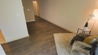 This single-floor space offers a straightforward and functional layout designed for practical living. The main living area is characterized by rich, dark hardwood-style flooring that runs consistently throughout, providing a warm and grounding aesthetic. The neutral beige walls complement the flooring, creating a cohesive and understated color palette that lends itself well to a variety of furnishing styles. White baseboards provide a crisp contrast along the perimeter of the room.

A plush, beige area rug anchors one corner of the living space, adding a soft textural element alongside a wicker accent piece. The room offers a generous open feel with ample wall space. A hallway extends toward the rear of the space, leading to additional rooms, including what appears to be a closet with louvered doors and a separate room beyond. The hallway maintains the same dark flooring, ensuring visual continuity throughout. Electrical outlets are conveniently positioned along the walls, and a light switch is visible near the hallway entrance. Overall, the space presents a clean, move-in-ready environment with a calm, neutral ambiance.