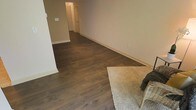 This single-floor space offers a straightforward and functional layout designed for practical living. The main living area is characterized by rich, dark hardwood-style flooring that runs consistently throughout, providing a warm and grounding aesthetic. The neutral beige walls complement the flooring, creating a cohesive and understated color palette that lends itself well to a variety of furnishing styles. White baseboards provide a crisp contrast along the perimeter of the room.

A plush, beige area rug anchors one corner of the living space, adding a soft textural element alongside a wicker accent piece. The room offers a generous open feel with ample wall space. A hallway extends toward the rear of the space, leading to additional rooms, including what appears to be a closet with louvered doors and a separate room beyond. The hallway maintains the same dark flooring, ensuring visual continuity throughout. Electrical outlets are conveniently positioned along the walls, and a light switch is visible near the hallway entrance. Overall, the space presents a clean, move-in-ready environment with a calm, neutral ambiance.