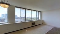 This single-floor space makes an immediate impression with its expansive wall of large, floor-to-ceiling windows that span the full width of the main living area. The windows offer unobstructed, panoramic views of a sprawling urban skyline, with city buildings, green spaces, and open sky stretching into the distance — a defining architectural feature that brings the outdoors in and floods the interior with natural light throughout the day. The elevated position of the unit enhances the sense of openness and scale, creating an airy, light-filled atmosphere. The living area features neutral-toned carpeting and clean white walls, providing a versatile, understated backdrop that allows the dramatic cityscape views to take center stage. A built-in climate control unit is integrated beneath the window line, maintaining comfort while preserving the sleek visual flow of the space. The minimalist interior design and open layout offer flexibility for a variety of furnishing configurations. Overall, the space combines functional simplicity with a standout architectural element — its commanding high-rise views — making it a distinctive and visually compelling environment.