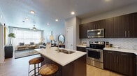 This thoughtfully designed single-floor residence offers a cohesive blend of modern finishes and functional living spaces. The open-concept kitchen is a standout feature, showcasing dark espresso cabinetry, white quartz countertops, a herringbone tile backsplash, and a full suite of stainless steel appliances including a range, microwave, and refrigerator. A central island with a double sink provides additional prep space and casual seating, while light wood-style flooring flows seamlessly into the adjoining dining and living areas. The dining space comfortably accommodates a full dining table and is enhanced by recessed lighting overhead. The living room is bright and welcoming, featuring large windows that invite abundant natural light, a ceiling fan, and generous space for comfortable seating arrangements. The bedroom is a calm retreat, carpeted for comfort, with a ceiling fan, wide windows, and ample room for a full furniture arrangement. The bathroom is well-equipped with dark wood cabinetry, a white countertop vanity, wood-look tile flooring, and a tiled shower, with direct access to a dedicated walk-in closet featuring built-in shelving. Throughout the residence, a neutral color palette and clean architectural lines create a polished, contemporary ambiance.