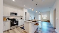 This well-designed one-bedroom, two-bathroom residence offers a thoughtfully laid-out single-floor plan with a bright, open-concept living space. The kitchen is a standout feature, showcasing white shaker cabinetry, granite countertops with a subway tile backsplash, stainless steel appliances including a range and built-in microwave, and a central island with bar seating — ideal for casual dining or entertaining. Pendant lighting above the island adds a contemporary touch, while recessed ceiling lights illuminate the space efficiently. Light hardwood-style flooring runs throughout, lending warmth and visual continuity. The kitchen opens seamlessly into the living area, which is equipped with a ceiling fan and benefits from natural light streaming through a large window and glass door that leads to an outdoor area. The bedroom is a comfortable, well-proportioned retreat with a ceiling fan, a window with natural light, and access to a dedicated closet with built-in shelving and hanging rods. A separate closet area with additional shelving provides further storage flexibility. The clean, neutral color palette of white walls and light flooring creates a bright, airy ambiance throughout the entire space.