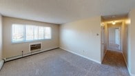 This single-floor unit offers a practical and straightforward layout, as illustrated by the 3D floor plan, which reveals a spacious living area, a kitchen, at least one bedroom, and a full bathroom arranged in a compact, efficient configuration.

The living area is carpeted and finished with neutral beige walls beneath a textured ceiling. A wide window fitted with horizontal blinds allows natural light to enter, while a wall-mounted air conditioning unit and baseboard heating ensure year-round climate control. An adjacent carpeted room, likely a bedroom, features a similar window, textured ceiling, and baseboard heating, maintaining a consistent aesthetic throughout.

The kitchen is efficiently organized, featuring dark wood upper cabinets on both sides, white laminate countertops, a stainless sink, a white refrigerator, a white electric range, and a black dishwasher. Wood-look flooring and a skylight or recessed overhead panel add a functional touch to the space.

The full bathroom is equipped with a single-sink vanity featuring a dark wood cabinet, a light-toned countertop, a wall-mounted mirror with built-in lighting, a white toilet, and a tiled shower-tub combination with a chrome showerhead and curtain rod. The overall ambiance of the unit is clean, functional, and move-in ready.