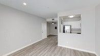 This single-floor residence offers a clean, open-concept layout that seamlessly connects the living area and kitchen. The living space is finished with light-toned hardwood-style flooring that runs throughout, complementing the neutral gray walls and bright white trim. Recessed lighting and a ceiling fan provide both functional illumination and air circulation, contributing to a comfortable atmosphere.

The kitchen is positioned adjacent to the main living area, separated by a half-wall peninsula that doubles as a breakfast bar. White cabinetry lines the upper portion of the kitchen, offering ample storage, while stainless steel appliances, including a full-size refrigerator, add a modern touch. The peninsula's raised countertop creates a natural division between the two spaces while maintaining an open, airy feel.

The entry door is visible from the living area, reinforcing the straightforward, accessible layout. Consistent flooring, neutral tones, and recessed lighting throughout give the space a cohesive and well-maintained appearance. Overall, the unit presents a practical and contemporary living environment suited for everyday comfort.
