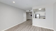 This single-floor residence offers a clean, open-concept layout that seamlessly connects the living area and kitchen. The living space is finished with light-toned hardwood-style flooring that runs throughout, complementing the neutral gray walls and bright white trim. Recessed lighting and a ceiling fan provide both functional illumination and air circulation, contributing to a comfortable atmosphere.

The kitchen is positioned adjacent to the main living area, separated by a half-wall peninsula that doubles as a breakfast bar. White cabinetry lines the upper portion of the kitchen, offering ample storage, while stainless steel appliances, including a full-size refrigerator, add a modern touch. The peninsula's raised countertop creates a natural division between the two spaces while maintaining an open, airy feel.

The entry door is visible from the living area, reinforcing the straightforward, accessible layout. Consistent flooring, neutral tones, and recessed lighting throughout give the space a cohesive and well-maintained appearance. Overall, the unit presents a practical and contemporary living environment suited for everyday comfort.