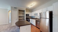 This single-floor residence offers a well-designed layout with distinct living zones connected by a cohesive neutral color palette and a mix of hardwood-style and carpeted flooring. The kitchen is a functional highlight, featuring white cabinetry, dark granite-style countertops, and a full suite of stainless steel appliances including a refrigerator, electric range, microwave, and dishwasher. A peninsula with a curved countertop extends into the adjacent space, providing additional prep area and casual seating. A built-in shelving nook adjacent to the kitchen offers convenient storage or workspace potential. The living area is generously sized, anchored by a white-framed fireplace and a sliding glass door that allows natural light to filter in through vertical blinds. Crown molding and a ceiling fan add architectural detail to this space. The bedroom is carpeted and bright, benefiting from a large window with outdoor views and a ceiling fan overhead. The bathroom includes a vanity with white cabinetry, dark countertops, a large mirror with overhead lighting, and a tub-shower combination with glass enclosure. Overall, the space presents a practical and comfortable layout with consistent finishes throughout.