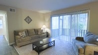 This single-floor residential space offers a comfortable and well-lit living environment designed for everyday functionality. The living room serves as the centerpiece of the space, featuring plush wall-to-wall carpeting that adds warmth underfoot. A defining feature of the room is the expansive floor-to-ceiling sliding glass door dressed with vertical blinds, which floods the interior with natural light and provides a seamless connection to the outdoors. The neutral, warm-toned walls create a clean and versatile backdrop, complementing the overall understated aesthetic of the space. The layout is open and accommodating, with ample room for seating and a central coffee table, making it well-suited for both relaxing and entertaining. Decorative wall art adds a touch of personality to the otherwise minimalist design. The single-floor layout ensures easy navigation throughout the space, promoting a cohesive and accessible flow from room to room. Overall, the space conveys a sense of calm simplicity, with its blend of natural light, neutral finishes, and practical layout making it a welcoming and livable environment.