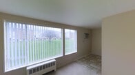This single-floor space presents a straightforward and functional layout finished in neutral tones throughout. The main living area is carpeted in a light beige, providing a soft underfoot feel, and is framed by smooth white baseboards and clean, understated walls. The space is open and versatile, lending itself well to a variety of furniture arrangements.

An adjacent area, likely a sunroom or secondary living space, features large sliding windows that span much of the wall, allowing a generous amount of natural light to fill the room. These windows offer views of a grassy outdoor area and surrounding buildings, creating a pleasant connection to the exterior. The flooring in this area transitions to a tile surface, adding a practical, easy-to-maintain finish. A wall-mounted air conditioning unit and a baseboard radiator are both present, providing year-round climate control.

A doorway visible in one area hints at additional rooms beyond, suggesting a connected and accessible floor plan. Overall, the space maintains a clean, move-in-ready aesthetic with simple finishes that offer a neutral canvas for personalization.