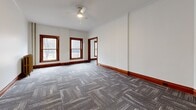 This well-appointed single-floor space showcases a thoughtful blend of classic architectural character and practical updates throughout its seven rooms. Rich hardwood door and window trim in warm walnut tones contrasts beautifully against crisp white walls, while patterned grey carpet tiles flow consistently across the main living areas, adding texture and a contemporary touch.

The layout offers a series of interconnected rooms, including a spacious main room bathed in natural light through multiple double-hung windows framed in matching wood trim. A ceiling fan overhead and original cast-iron radiators speak to the building's historic roots while providing functional comfort. Crown molding adds an elegant finishing detail to several rooms.

The kitchen has been thoughtfully updated with white shaker-style cabinetry, light countertops, a stainless steel sink, a gas range, and a dishwasher, all set on classic tile flooring. The bathroom retains its vintage charm with a clawfoot soaking tub, classic black-and-white mosaic tile flooring, white subway wall tiles, and a traditional window that brings in natural light. Solid wood panel doors with original hardware complete the period aesthetic, making this a distinctive space that honors its architectural heritage.