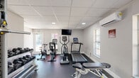 Located at 5528 Cinderlane Pkwy in Orlando, FL, this fitness center is a dedicated single-room workout facility designed to support a well-rounded exercise routine. The space features a clean, modern aesthetic with light gray walls, white baseboards, and a drop tile ceiling equipped with recessed panel lighting that keeps the room bright and functional throughout the day.

Multiple windows along the walls allow natural light to fill the space while offering views of the surrounding outdoor area. A wall-mounted split air conditioning unit ensures comfortable climate control year-round. The durable rubber flooring provides traction and cushioning appropriate for a variety of workout activities.

The cardio section is outfitted with a treadmill, elliptical trainer, and stationary spin bike, all positioned near the windows for an airy feel. A wall-mounted flat-screen television is positioned for easy viewing during cardio sessions. The strength training area includes a cable cross machine, a dedicated leg press or curl machine, a multi-tiered dumbbell rack with a full set of free weights, and an adjustable workout bench. A sanitation station with a paper towel dispenser is mounted on the wall for hygiene convenience. Two entry doors provide easy access to and from the facility, making this fitness center a practical and comprehensive amenity.