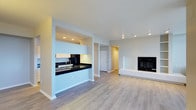 Unit 3702 is a single-floor residence designed with an open, flowing layout that maximizes the sense of space. The living area is anchored by a sleek, built-in fireplace framed by a clean white surround, creating a focal point that adds warmth and character to the room. Light wood-style flooring runs throughout the unit, lending a modern yet inviting aesthetic that ties the spaces together seamlessly.

The kitchen is positioned behind a pass-through counter with dark granite or quartz countertops that contrast beautifully against light cabinetry above. This peninsula-style opening connects the kitchen to the main living area, making it ideal for entertaining or casual dining. Recessed lighting in the kitchen provides focused illumination, complementing the natural light that filters into the broader living space.

Neutral gray-toned walls throughout the unit create a versatile backdrop, allowing for a wide range of interior design styles. The open corridor leading toward additional rooms is lined with closet storage, suggesting practical functionality alongside the clean aesthetic. Overall, the unit presents a contemporary, well-appointed living environment with thoughtful design details throughout.