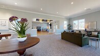 This single-story community clubhouse is thoughtfully designed to provide a welcoming and functional gathering space. The entry foyer greets visitors with gleaming hardwood floors, a coffered ceiling with an elegant pendant light, and glass double doors that frame views of the surrounding neighborhood. Crown molding and soft gray walls establish a clean, contemporary aesthetic carried throughout the facility.

The main lounge area transitions between carpeted and hardwood zones, accommodating multiple seating arrangements with plush sofas and accent chairs. A dramatic stone fireplace anchors one end of the room, rising toward soaring ceilings with clerestory windows that flood the space with natural light. A wall-mounted television above the fireplace adds a modern amenity.

An open-plan kitchen area features warm wood cabinetry, white quartz countertops, a subway tile backsplash, floating black shelves, and a bold blue island, with direct glass door access to a covered outdoor patio supported by stone columns.

A dedicated leasing or reception area includes carpeted floors, recessed lighting, a central display table, and community information displayed along the walls. Throughout, generous windows provide views of the surrounding development, reinforcing a bright and connected atmosphere.