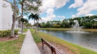 This tranquil single-level space is defined by its picturesque lakefront setting, characteristic of South Florida's natural landscape. A gently curving paved walkway runs alongside the water's edge, lined with towering palm trees and mature shade trees that frame the views beautifully. A classic wooden bench positioned along the path invites visitors to pause and take in the peaceful surroundings.

The centerpiece of the outdoor area is a well-maintained pond, its calm surface reflecting the surrounding greenery and the expansive blue sky above. A decorative water fountain rises from the center of the lake, adding both visual interest and a soothing ambient sound to the environment. Manicured green lawns extend along the water's edge, creating a clean and open feel throughout.

The lush tree canopy — a mix of palms, oaks, and tropical species — provides both shade and a sense of privacy, while the open sky above allows for abundant natural light. Views across the lake reveal additional greenery and neighboring structures in the distance, contributing to a sense of community and spaciousness. This well-kept outdoor setting offers a calm, park-like atmosphere well suited for leisurely walks, relaxation, and enjoyment of the natural environment.
