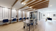 Located in Oviedo, FL, this thoughtfully designed single-floor amenity space offers a versatile and visually engaging environment for collaboration and productivity. The interior is anchored by a central conference table surrounded by modern seating, providing an ideal setting for group meetings or social gatherings. Flanking the main area, a row of sleek individual workstations lines the walls, each accented by literary-themed wall graphics that add a distinctive, cultured character to the space.

Architectural interest is introduced through striking diagonal wood beam detailing along the ceiling, which adds warmth and visual depth against the crisp white surfaces. The flooring transitions between light wood-plank and geometric hexagonal tile patterns, creating distinct yet cohesive zones throughout the layout. A series of glass-paneled private rooms with black metal frames line one side of the space, offering enclosed areas for focused work or intimate meetings.

Natural light filters in through French doors at one end, which open to an outdoor balcony view, further brightening the already light-filled interior. A bold fireplace canvas artwork anchors one wall, adding warmth and personality. Curated modern art pieces and navy blue furnishings with gold accents complete the polished, contemporary aesthetic, making this a functional and inspiring communal destination.