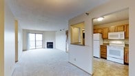 This single-floor residence is designed with an open-concept layout that seamlessly connects the main living area to the kitchen. The spacious living room is finished with plush carpet and features a classic fireplace as a central focal point, adding both warmth and character to the space. Large sliding glass doors flood the room with natural light and provide direct access to a private balcony, offering outdoor views and ventilation.

A partial-height dividing wall separates the living area from the kitchen while maintaining an open, airy feel throughout. The kitchen is equipped with warm-toned wood cabinetry offering generous storage, a white refrigerator, and light-colored flooring that contrasts neatly with the carpeted living space. A pass-through window or opening connects the kitchen visually to the adjacent hallway area, enhancing the sense of flow. The textured ceiling adds a subtle design detail throughout the space. Overall, the layout is practical and well-organized, with a neutral color palette that creates a bright and welcoming ambiance.