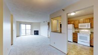 This single-floor residence is designed with an open-concept layout that seamlessly connects the main living area to the kitchen. The spacious living room is finished with plush carpet and features a classic fireplace as a central focal point, adding both warmth and character to the space. Large sliding glass doors flood the room with natural light and provide direct access to a private balcony, offering outdoor views and ventilation.

A partial-height dividing wall separates the living area from the kitchen while maintaining an open, airy feel throughout. The kitchen is equipped with warm-toned wood cabinetry offering generous storage, a white refrigerator, and light-colored flooring that contrasts neatly with the carpeted living space. A pass-through window or opening connects the kitchen visually to the adjacent hallway area, enhancing the sense of flow. The textured ceiling adds a subtle design detail throughout the space. Overall, the layout is practical and well-organized, with a neutral color palette that creates a bright and welcoming ambiance.