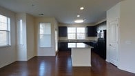 This single-story space showcases a well-appointed kitchen that serves as the focal point of the layout. Dark espresso-toned cabinetry lines the walls, providing a striking contrast against the light-colored walls and cream-toned granite countertops. A central island with a white base and matching granite surface offers additional workspace and a natural gathering point within the kitchen.

The flooring throughout is rich, dark hardwood, adding warmth and a sense of continuity to the space. Recessed lighting and a flush-mount ceiling fixture ensure even illumination, while multiple windows fitted with white horizontal blinds allow ample natural light to filter in, brightening the interior considerably.

Black appliances, including a refrigerator and range, complement the dark cabinetry for a cohesive, modern aesthetic. The kitchen layout follows a practical U-shaped design along the perimeter walls, maximizing storage and counter space. The open feel of the area, combined with the interplay of dark and light tones, creates a functional yet visually balanced environment well-suited for both everyday living and entertaining.