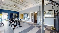 This fitness center is thoughtfully designed to provide a comprehensive and comfortable workout environment. The space is defined by its striking coffered ceiling with recessed panels and ceiling fans, lending an elevated, architectural character uncommon in typical gym facilities. The flooring features a two-tone checkered pattern of light tile and rubberized gray material, offering both durability and a polished aesthetic.

Along one wall, a row of spin bikes is neatly arranged, ideal for cycling workouts, while additional cardio equipment, including treadmills, is visible along the mirrored accent wall. The mirrors, framed in white and set against a bold navy blue wall, serve to visually expand the space and provide users with proper form feedback during exercise.

Framed artwork adds a decorative touch between the mirrors, enhancing the overall ambiance. A separate room is accessible through glass-paneled doors, suggesting a dedicated area for additional equipment or studio-style classes. Neutral gray walls, recessed lighting, and ceiling fans contribute to a clean, modern atmosphere that balances functionality with refined design throughout the space.