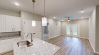 Located at 143 Emerson Point Dr in Burlington, NC, this single-floor, 2-bedroom, 2-bathroom residence offers a well-designed layout with a clean, modern aesthetic throughout. The entrance leads down a bright hallway with multiple closets, opening into a combined dining, kitchen, and living area that serves as the heart of the home. The kitchen is a standout feature, boasting crisp white shaker cabinetry, granite countertops, a center island with an undermount sink and pendant lighting, and stainless steel appliances. Light wood-look flooring runs throughout the main living areas, complementing the neutral, warm color palette. The open living room is filled with natural light and is anchored by a ceiling fan, with French doors providing outdoor access and views of surrounding greenery. A dedicated pantry and in-unit laundry add everyday convenience. Both bedrooms are carpeted for comfort, each offering a calm, neutral atmosphere and access to closet space. The bathrooms mirror the kitchen's design language, featuring granite-topped white vanities and wood-look flooring. A covered exterior entry with a pergola-style structure completes this thoughtfully appointed residence.