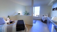 This single-floor space presents a clean, minimalist design with a calming ambiance well-suited for wellness or therapeutic use. The open layout is anchored by light wood flooring that extends throughout, contributing to a warm yet understated atmosphere. A partial dividing wall creates a subtle separation between areas without fully closing off the space, maintaining an airy, open feel while offering a degree of privacy.

Natural light is a defining feature, with a large window allowing bright, diffused light to fill the space and provide views to the exterior. The window ledge serves as a display area for greenery, including a lush fern and smaller potted plants, adding life and a natural element to the interior. Candles and minimal decorative objects complement the tranquil tone.

The treatment or relaxation area is furnished with a padded table dressed in a striped covering and topped with a tufted bench, suggesting use as a massage or bodywork room. A draped service table nearby supports the functional nature of the space. A yoga mat positioned near the window further reinforces the wellness-oriented purpose of this thoughtfully arranged, light-filled environment.