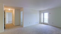 This single-floor space presents a clean, open layout finished in neutral white tones throughout. The main living area is generously sized, featuring plush wall-to-wall carpeting that adds warmth and comfort underfoot. White walls and standard-height ceilings contribute to a bright, airy atmosphere, further enhanced by a glass-paneled exterior door along one wall that allows natural light to filter into the space and provides access to the outdoors. A baseboard heating unit is positioned beneath the exterior door, ensuring efficient climate control. An open doorway on the opposite side of the room leads into an adjoining hallway or additional room, offering a glimpse of further living space beyond. The corridor appears to connect to additional rooms, suggesting a functional and practical floor plan. Overall, the space maintains a simple, neutral aesthetic with consistent flooring and a clean finish throughout, providing a versatile blank canvas well-suited for a variety of furnishing arrangements and personal styles.