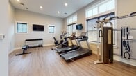 Located at 5833 Evans Farm Dr. in Lewis Center, Ohio, this fitness center offers a well-appointed, single-floor exercise facility designed to support a comprehensive workout experience. The space features durable light wood-tone flooring throughout, complemented by neutral gray walls and clean white trim that create a bright, modern aesthetic. Recessed ceiling lighting ensures consistent illumination across the entire area.

The cardio zone is equipped with multiple Spirit-brand treadmills and an elliptical trainer, positioned alongside large windows dressed with horizontal blinds that allow natural light to fill the room. Two wall-mounted flat-screen TVs are strategically placed for viewing during workouts. A cable resistance machine and a recumbent bike add further variety to the cardio and strength training options available.

The free weight area features a full dumbbell rack stocked with a range of weights, accompanied by a dedicated workout bench for strength training exercises. Stability and exercise balls are also present, supporting flexibility and functional fitness routines. The space is well-organized with room to move freely between equipment.

Two interior doors provide access to adjacent areas, including what appears to be a connected common or lounge space visible through a glass-paned door. The overall environment is clean, functional, and welcoming, making this fitness center a practical and attractive amenity for residents of the Evans Farm community.