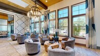 This single-story amenity space is designed with a contemporary aesthetic that balances elegance and functionality across several well-defined areas. The centerpiece lounge features soaring ceilings with warm wood beam accents and a striking multi-tiered chandelier with exposed bulbs, casting a warm glow over a curated seating arrangement of plush gray swivel chairs surrounding a geometric wood coffee table. Floor-to-ceiling windows with teal and white drapery flood this area with natural light and offer views of lush outdoor greenery and palm trees.

Adjacent to the lounge, a long white quartz bar counter with leather barstools serves as a social gathering point, complemented by teal glass tile backsplash, pendant lighting, and a built-in beverage station. A banquette seating area with marble-top tables and bold geometric wall patterning provides an intimate setting for casual meetings or dining.

The space also includes glass-walled private workrooms with carpeted floors and ample natural light, offering quiet, focused environments. A recreation area features a shuffleboard table alongside large windows overlooking an outdoor patio. Patterned geometric wall art and designer accent walls unify the interior's cohesive, upscale style throughout.