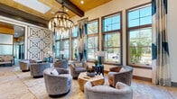 This single-story amenity space is designed with a contemporary aesthetic that balances elegance and functionality across several well-defined areas. The centerpiece lounge features soaring ceilings with warm wood beam accents and a striking multi-tiered chandelier with exposed bulbs, casting a warm glow over a curated seating arrangement of plush gray swivel chairs surrounding a geometric wood coffee table. Floor-to-ceiling windows with teal and white drapery flood this area with natural light and offer views of lush outdoor greenery and palm trees.

Adjacent to the lounge, a long white quartz bar counter with leather barstools serves as a social gathering point, complemented by teal glass tile backsplash, pendant lighting, and a built-in beverage station. A banquette seating area with marble-top tables and bold geometric wall patterning provides an intimate setting for casual meetings or dining.

The space also includes glass-walled private workrooms with carpeted floors and ample natural light, offering quiet, focused environments. A recreation area features a shuffleboard table alongside large windows overlooking an outdoor patio. Patterned geometric wall art and designer accent walls unify the interior's cohesive, upscale style throughout.