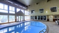 This single-floor indoor pool facility offers a striking and well-appointed aquatic environment designed for comfort and year-round use. The centerpiece is a large, freeform swimming pool with smooth, curving edges and clear blue water, framed by a spacious surrounding deck. The space is defined by its dramatic double-height ceilings clad in warm wood-toned paneling, which lend the area a welcoming, lodge-like character. Towering angled windows along one wall flood the interior with abundant natural light, creating an airy, open atmosphere and providing views of the surrounding greenery outside. Entry doors along the windowed wall offer convenient access to an outdoor area. An elevated interior overlook, visible through large glass windows on the opposing wall, provides a view into an adjacent space — suggesting a fitness center or lounge area — and adds a layered, multi-functional dimension to the facility. Exposed ductwork integrated into the ceiling reflects a practical yet thoughtfully designed mechanical layout. Together, the architectural details, generous natural lighting, and the inviting pool make this a standout recreational amenity suited for leisure and fitness alike.