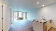 Located at 910 SW 18th Ave in Portland, Oregon, this single-floor unit offers a contemporary design with clean lines, light-toned hardwood-style flooring, and a bright, neutral color palette throughout. The open-concept living and kitchen area serves as the heart of the space, where natural light filters in through a bank of large windows offering views of the surrounding urban streetscape, including glimpses of mature trees and the Portland skyline.

The kitchen is well-appointed with stainless steel appliances, including a gas range, a French door refrigerator, and a built-in microwave. Light wood cabinetry extends to the ceiling, complemented by a geometric tile backsplash and a peninsula with an undermount sink — ideal for casual dining or meal preparation. Recessed lighting provides additional illumination throughout.

The living area is generously proportioned, with a wall-mounted PTAC heating and cooling unit positioned beneath the windows for year-round climate control. A separate bedroom features tall, narrow windows with urban views and warm hardwood-style flooring. The bathroom is finished with a wood-tone vanity, a white undermount sink, and a tub-shower combination with white subway tile surround.