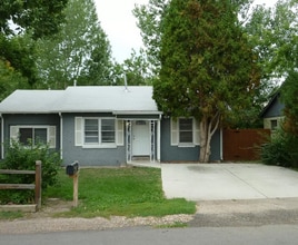 Quaint single-story gray house with a white roof, front porch, and lush green surroundings. - 1385 Yukon Street