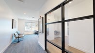 Located at 2005 Brook Rd in Richmond, Virginia, this contemporary single-floor hallway space delivers a polished, functional environment with a refined modern aesthetic. The layout flows seamlessly from entry to workstation, finished with soft blue-grey carpeting underfoot and crisp white walls that create a clean, professional atmosphere.

A standout design feature is the bold black-framed glass partition wall, which visually connects the hallway to an adjacent tiled restroom area while maintaining a sense of openness and transparency. Natural light filters in through a large window at the far end, illuminating the space and offering views of the surrounding exterior.

A built-in long white desk runs along one wall, accented by a textured stone tile feature wall and paired with warm brass gooseneck sconce lighting — lending an upscale, design-forward touch. Ergonomic task chairs and casual seating options are thoughtfully positioned throughout, supporting both focused work and relaxed interaction.

Black-framed glass entry doors echo the interior partitions, reinforcing a cohesive industrial-modern design language. Recessed ceiling lighting provides consistent, even illumination throughout. The overall ambiance is bright, clean, and purposefully designed — striking a balance between contemporary style and practical functionality suited for a professional setting.