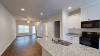 This two-story space offers a well-designed open-concept main level that seamlessly connects the kitchen and living areas. The kitchen is a standout feature, showcasing granite countertops with a speckled finish, a double stainless steel sink, and crisp white cabinetry that extends to the upper walls, providing ample storage. Recessed lighting illuminates the kitchen area, complementing the natural light that filters in through windows in the adjacent living space.

The living area extends comfortably from the kitchen, featuring warm hardwood flooring that runs throughout the main level. A ceiling fan with a light fixture serves as a functional centerpiece in the living area, while large windows along one wall invite generous natural light. Neutral-toned walls contribute to a clean, airy atmosphere throughout.

The main level also includes access to what appears to be a closet or storage space, as well as a staircase leading to the second floor. The open layout between the kitchen and living area creates a cohesive flow, making the space well-suited for both everyday living and entertaining. The overall design balances practicality with modern aesthetics.