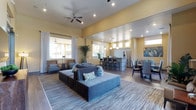 This single-floor space showcases an open-plan layout that seamlessly connects multiple functional zones. The main lounge area is anchored by a large, low-profile sectional sofa in cool gray tones, accented with decorative pillows and positioned atop a wide striped area rug. Ceiling fans with recessed lighting overhead contribute to both comfort and a bright, airy atmosphere. Warm yellow walls lend a welcoming and energetic tone throughout the space.

Large windows dressed with floor-length drapes allow generous natural light to pour in, further enhancing the open feel. Adjacent to the lounge, a kitchen and bar area features a raised counter with bar-height seating, pendant lighting, and stainless steel appliances, making it well-suited for gatherings or events. A formal dining area with upholstered chairs sits nearby, offering a more structured seating option.

Accent seating near the windows provides a quieter retreat within the larger space, while indoor greenery adds a natural, refreshing element. Hardwood-style flooring ties the zones together cohesively, creating a polished, contemporary aesthetic ideal for communal or social use.