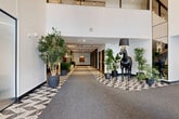 Located at 2600 Boulevard Cavendish in Montréal, this expansive lobby complex combines a fitness facility, coworking lounge, and grand entrance hall into a single cohesive, thoughtfully designed floor. The main corridor makes an immediate impression with its double-height ceilings, bold geometric-patterned flooring, lush potted greenery, and a dramatic life-sized black horse sculpture. Track lighting runs the length of the hallway, drawing the eye toward curated artwork at its end. The entrance hall features soaring floor-to-ceiling glazing, exposed concrete columns, and elegant globe chandeliers, creating a bright, welcoming arrival experience. The fitness center is well-appointed with cardio machines, weight benches, and free weights, wrapped in large windows that frame outdoor views and flood the space with natural light. Full-length mirrors and distinctive striped columns add a polished, design-forward aesthetic. The lounge area offers a warm contrast, with a coffered wood-paneled ceiling, a fireplace flanked by teal millwork, a plush navy velvet sofa, and generous glazing overlooking the streetscape. A dedicated coworking zone features glass-partitioned offices, light wood flooring, and modern furnishings, providing a professional yet comfortable work environment throughout.