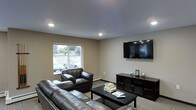This single-floor space features a well-appointed living area designed for comfort and functionality. The room is finished with neutral beige walls and a smooth gray ceiling equipped with recessed lighting, creating a clean and inviting atmosphere. A large window faces the street, allowing natural light to fill the space while offering views of the surrounding neighborhood.

The seating area is furnished with dark brown leather sofas and a matching armchair arranged around a black rectangular coffee table, which holds a selection of reading materials. A wall-mounted flat-screen television serves as a focal point on one side of the room, positioned above a dark wood media console that provides additional storage. The console is accessorized with small decorative items, adding a subtle personal touch.

The flooring consists of a patterned area rug layered over what appears to be neutral-toned carpeting, contributing to a warm and grounded feel. A baseboard heating unit runs along the wall beneath the window, indicating climate control capabilities. Overall, the space presents a functional and comfortable layout suited for relaxation and everyday use.