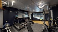 This fitness center offers a comprehensive workout environment designed with a modern industrial aesthetic. The space is divided into two distinct flooring zones: textured carpet tiles cover the primary exercise area, while smooth hardwood flooring defines a dedicated functional training and stretching zone near the windowed perimeter, allowing natural light to filter in.

The cardio section houses multiple True-brand treadmills and elliptical machines, each equipped with individual SKY digital display screens. Large floor-to-ceiling mirrors line the walls throughout, visually expanding the space and allowing users to monitor their form during workouts. The strength training area features a Hoist multi-functional cable machine, a set of spin bikes, and a well-organized dumbbell rack offering a range of weights.

Exposed ductwork, colorful overhead pipes in orange and yellow tones, and recessed lighting contribute to the space's industrial character. A wall-mounted dual water fountain station with an adjacent hand sanitizer dispenser provides convenience. The ceiling features a mix of drop tile and open industrial elements, reinforcing the modern-utilitarian design. A glass-paneled door connects the fitness area to an adjacent room, enhancing accessibility throughout the facility.