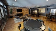 This single-floor entertainment lounge is designed for socializing and recreation, combining industrial charm with sophisticated style. The space is anchored by a striking exposed brick accent wall that adds warmth and texture, creating a compelling visual backdrop for the wall-mounted flat-screen television and dark wood media console beneath it. A curated seating area nearby features leather lounge chairs arranged around a low coffee table atop a neutral area rug, offering a comfortable spot for casual conversation or watching TV. At the forefront of the room sits a dedicated poker table surrounded by cushioned gaming chairs, ideal for hosting card nights. Toward the rear, a billiards table takes center stage beneath a sleek pendant light fixture, providing the perfect setting for friendly competition. The hardwood flooring flows throughout, unifying the various zones of the space. Recessed lighting in the ceiling complements the pendant fixtures to create a warm and inviting ambiance. A pass-through wet bar or service window is visible along one wall, adding convenience for entertaining. Dark wall accents and thoughtful furnishing choices throughout give this lounge a polished, upscale feel well-suited for both relaxed gatherings and lively events.