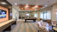 This single-floor community lounge and recreation room is thoughtfully designed to offer both entertainment and relaxation. The space is anchored by a striking coffered ceiling detail finished in a warm copper-orange tone, framed by recessed lighting that casts a welcoming glow throughout the room. Pendant lights hang above the central pool table, adding a focused ambiance to the gaming area.

The layout accommodates a variety of activities, with a full-size pool table as the centerpiece and a shuffleboard table positioned along one wall. A dedicated media area features a wall-mounted flat-screen television, flanked by pendant lighting and a casual seating arrangement, making it ideal for group viewing. A dining or activity table with bar-height seating occupies a defined nook nearby.

Plush, patterned accent chairs in coral and grey tones provide comfortable seating along the perimeter, adding a contemporary design touch. Natural light filters through large windows dressed with sheer curtains, brightening the space. The flooring features a combination of wood-look planks and carpeted sections, adding texture and visual depth to the overall design.