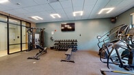 This contemporary fitness center is thoughtfully designed to support a full range of workout routines across multiple distinct zones. The space features soft teal-toned walls, a drop ceiling with recessed linear lighting, and durable carpeted flooring throughout the main exercise areas, creating a clean and energizing atmosphere.

The strength training area is well-appointed with a multi-functional cable machine, a full rack of dumbbells ranging across various weights, and adjustable workout benches — all anchored beneath wall-mounted flat-screen televisions. An adjacent cardio zone houses stair climbers and cycling equipment, with large windows that draw in natural light and offer views of the surrounding exterior.

Floor-to-ceiling mirrors line one wall of the open exercise floor, visually expanding the space and supporting functional training and stretching activities. A dedicated turf zone adds versatility, equipped with a rowing machine, plyo box, a functional training rig, and a striking floor-to-ceiling forest mural that adds a dynamic, nature-inspired focal point to the room.

The facility also includes a transitional corridor finished with elegant herringbone-patterned hardwood flooring, black-framed glass partitions, and access to restroom facilities and a water station, ensuring a complete and polished experience for users.