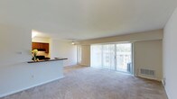 This single-floor space offers a clean, open layout that connects the kitchen and living area in a seamless, functional design. The living area is finished with wall-to-wall carpeting in a neutral tone, creating a warm and inviting atmosphere throughout. A ceiling fan is centrally positioned overhead, providing both practical air circulation and a subtle design accent.

The kitchen is situated adjacent to the living space and is defined by rich, warm-toned wood cabinetry paired with a dark countertop that extends to form a breakfast bar or pass-through — an ideal feature for casual dining or entertaining. A cooktop and range hood are visible, indicating a fully functional cooking area.

Natural light is a standout feature of this space. A large sliding glass door, dressed with vertical blinds, spans much of the exterior wall and opens to an outdoor area, flooding the interior with daylight and providing easy access to the outside. An additional window further enhances the bright, airy feel of the room.

The walls are finished in a soft, neutral tone that complements the light-filled environment and contributes to the overall sense of openness. The layout is straightforward and versatile, making it well-suited for a variety of furniture arrangements and lifestyle needs.