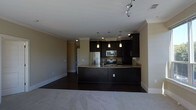 This single-floor space is designed with an open-concept layout that seamlessly connects the kitchen and living area, creating a cohesive and functional environment. The kitchen is a standout feature, outfitted with dark espresso cabinetry that extends to the upper walls, offering generous storage capacity. The cabinetry is complemented by light stone countertops that wrap around a curved breakfast bar, providing both workspace and casual seating. Stainless steel appliances, including a refrigerator, microwave, and range, are integrated into the design for a polished, contemporary look. Three pendant lights hang above the kitchen area, providing focused task lighting, while recessed lighting and a track lighting system illuminate the broader space. Crown molding lines the ceiling throughout, adding a refined architectural detail. The living area features plush carpeting in a neutral tone, contrasting with the dark hardwood-style flooring in the kitchen. The overall color palette — warm beige walls, dark cabinetry, and light countertops — creates a balanced, inviting ambiance suited to modern living.