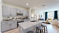 This thoughtfully designed single-floor residence combines modern finishes with a functional, open layout. The kitchen serves as a central highlight, showcasing gray shaker-style cabinetry with matte black hardware, granite-look countertops, a large center island with bar seating, and a full suite of stainless steel appliances including a French door refrigerator, electric range with microwave, all set atop light-toned hardwood-style flooring. The kitchen flows seamlessly into the living area, where large windows dressed with floor-to-ceiling draping invite generous natural light and create a bright, welcoming atmosphere. The bedrooms are generously sized, featuring plush carpeting, high ceilings, and windows that bring in soft natural light. Each bedroom connects to a well-finished bathroom, complete with granite-topped gray vanities, subway tile shower surrounds, and light hardwood-style flooring. Spacious walk-in closets accompany the bedrooms, offering ample built-in shelving and hanging storage. The overall aesthetic throughout the residence is clean and contemporary, with a cohesive neutral palette of whites, grays, and warm wood tones that lend the space a polished yet inviting character.