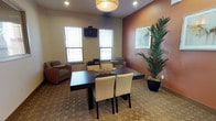This single-floor communal space is designed to serve as a gathering and meeting area, combining functionality with a warm, contemporary aesthetic. The main room is anchored by a dark espresso dining table surrounded by cream upholstered chairs, positioned beneath a woven drum pendant light that adds a decorative focal point overhead. Recessed lighting throughout ensures the space is well-lit, complementing the natural light admitted by multiple windows fitted with white blinds.

A standout design feature is the rich terracotta-toned accent wall, which introduces warmth and depth to the otherwise neutral palette of beige and white. Framed artwork adorns this wall alongside a tall potted palm, adding a natural, organic element to the interior. Comfortable lounge chairs are positioned near the windows, creating a relaxed seating area ideal for informal gatherings.

A wall-mounted flat-screen television is integrated into the space, enhancing its utility for presentations or entertainment. Large interior pass-through windows connect the main area to an adjacent kitchenette, visible through both the windows and a glazed panel door. The patterned tile flooring runs consistently throughout, grounding the space with texture. Overall, the room strikes a balance between casual comfort and practical functionality.